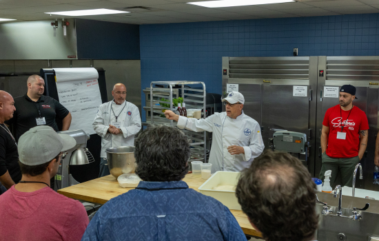 Chef in white coat instructing class participants gathered around in counter in a professional back-of- house kitchen setting